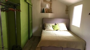 Un dormitorio con una cama, una pared verde, suelo de madera, una ventana y una lámpara sobre una estantería.