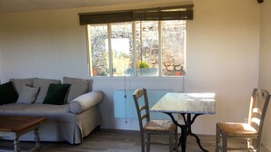 Una habitación acogedora con sofá, una mesa y sillas. Se ve una ventana con vistas a un muro de piedra y plantas.
