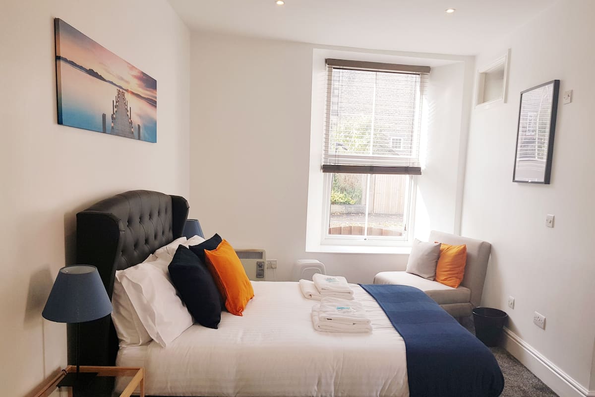 A hotel room with a bed, a bedside table, a lamp, a window with blinds, a chair, and a painting on the wall.