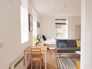 A modern living room with a sofa, a dining table with chairs, a small round table, and a window with blinds. - BlueOne Serviced Apartments-Sandon House (Sheffield)