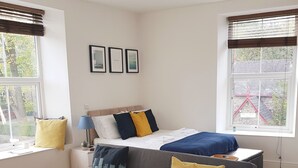Une chambre moderne avec un lit, un canapé gris, une petite table et une fenêtre avec des stores.