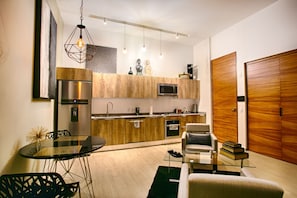 A modern kitchen with a built-in microwave, oven, and stainless steel refrigerator. There is a dining area with a glass table and chairs, and a wooden door in the background.