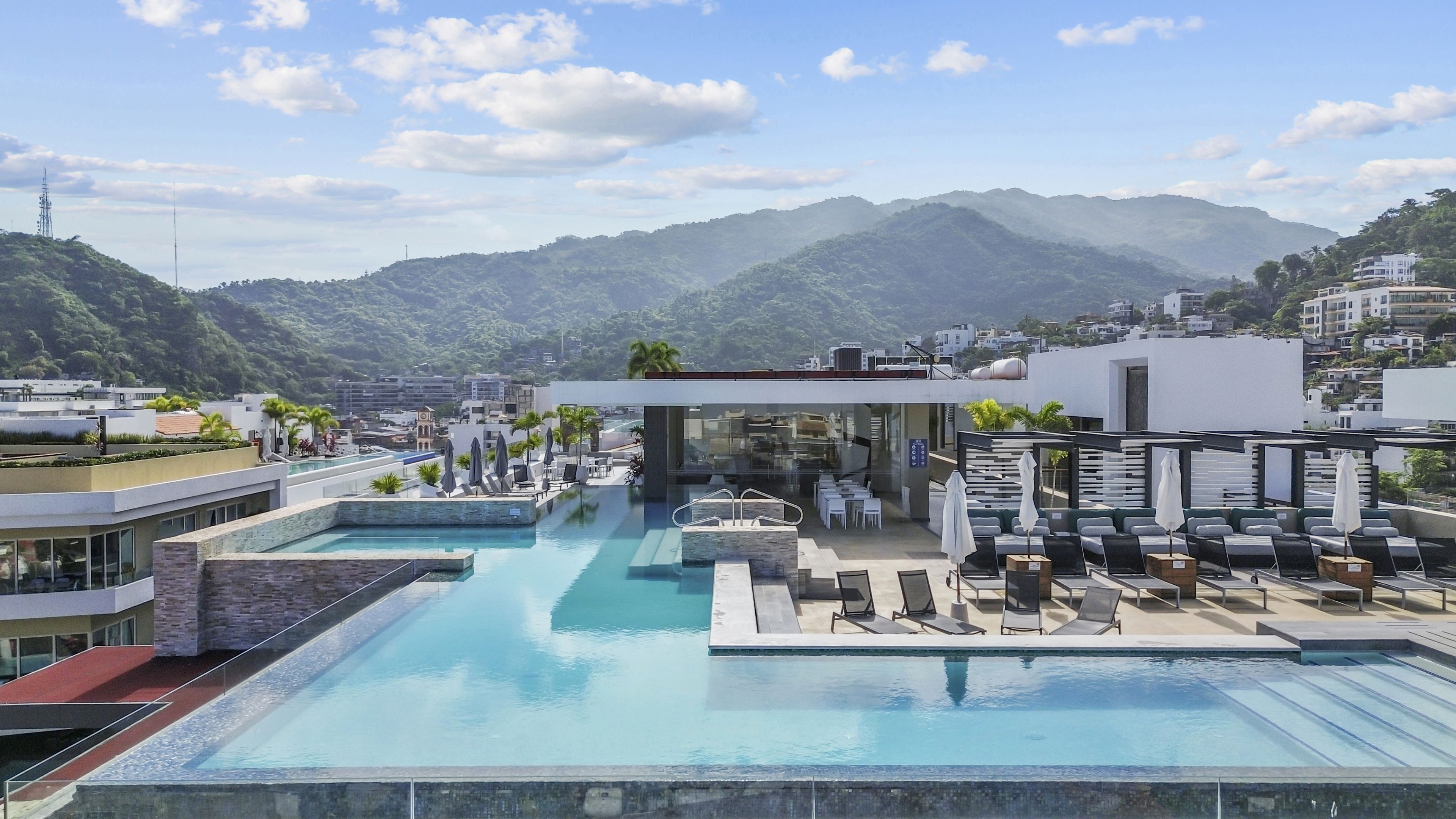 A rooftop infinity pool with lounge chairs and a dining area, overlooking a mountainous landscape.