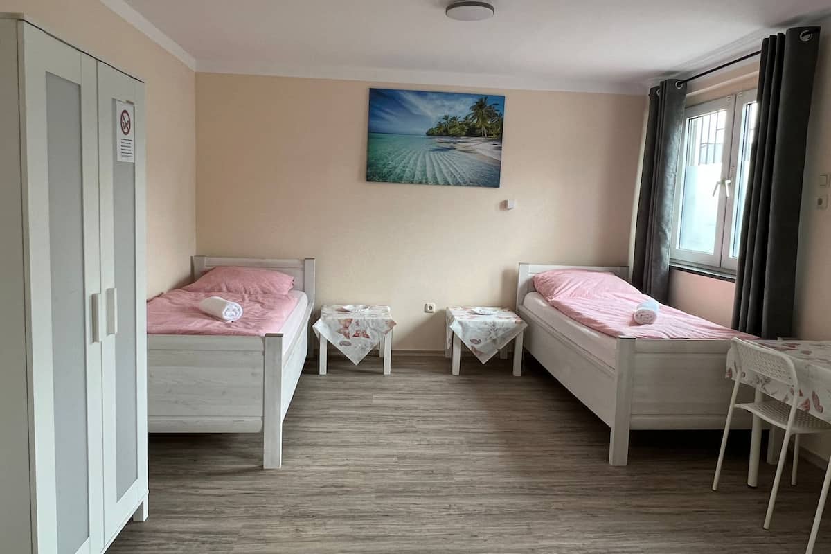 A room with two beds, a table, a chair, a wardrobe, and a painting on the wall.