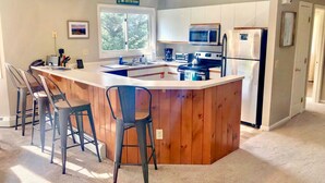 Una cocina moderna con una isla de madera, taburetes de bar, un microondas, un refrigerador y un ventanal que proporciona luz natural.