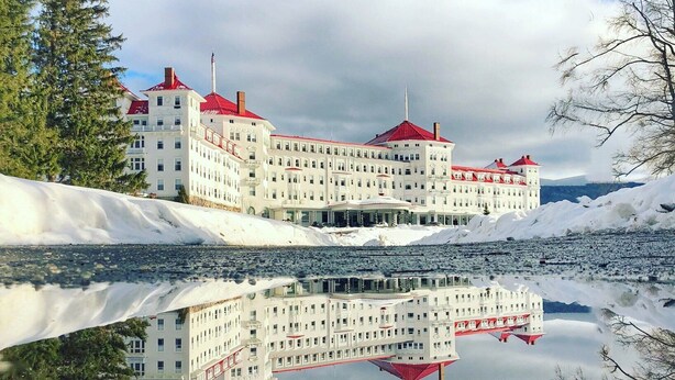 Un hotel grande y de varios pisos con techo rojo, ubicado junto a un cuerpo de agua tranquilo que refleja el edificio.