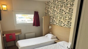 A compact room with two single beds, a small desk, a chair, a radiator, a window with curtains, and a wall with a leaf pattern. - Hôtel Classe Eco Reims Tinqueux (Tinqueux)