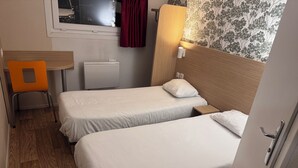 A small hotel room with two single beds, a wooden desk, a chair, a radiator, and a window with a curtain. - Hôtel Classe Eco Reims Tinqueux (Tinqueux)