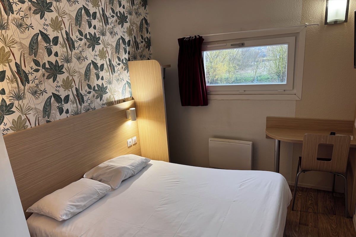A hotel room with a bed, a wooden headboard, a window with a view, a small table, and a radiator.