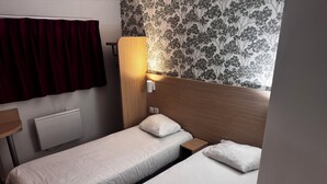 A hotel room with two single beds, a wooden headboard, a small bedside table, a wall-mounted lamp, a radiator, and a window with a curtain. - Hôtel Classe Eco Reims Tinqueux (Tinqueux)