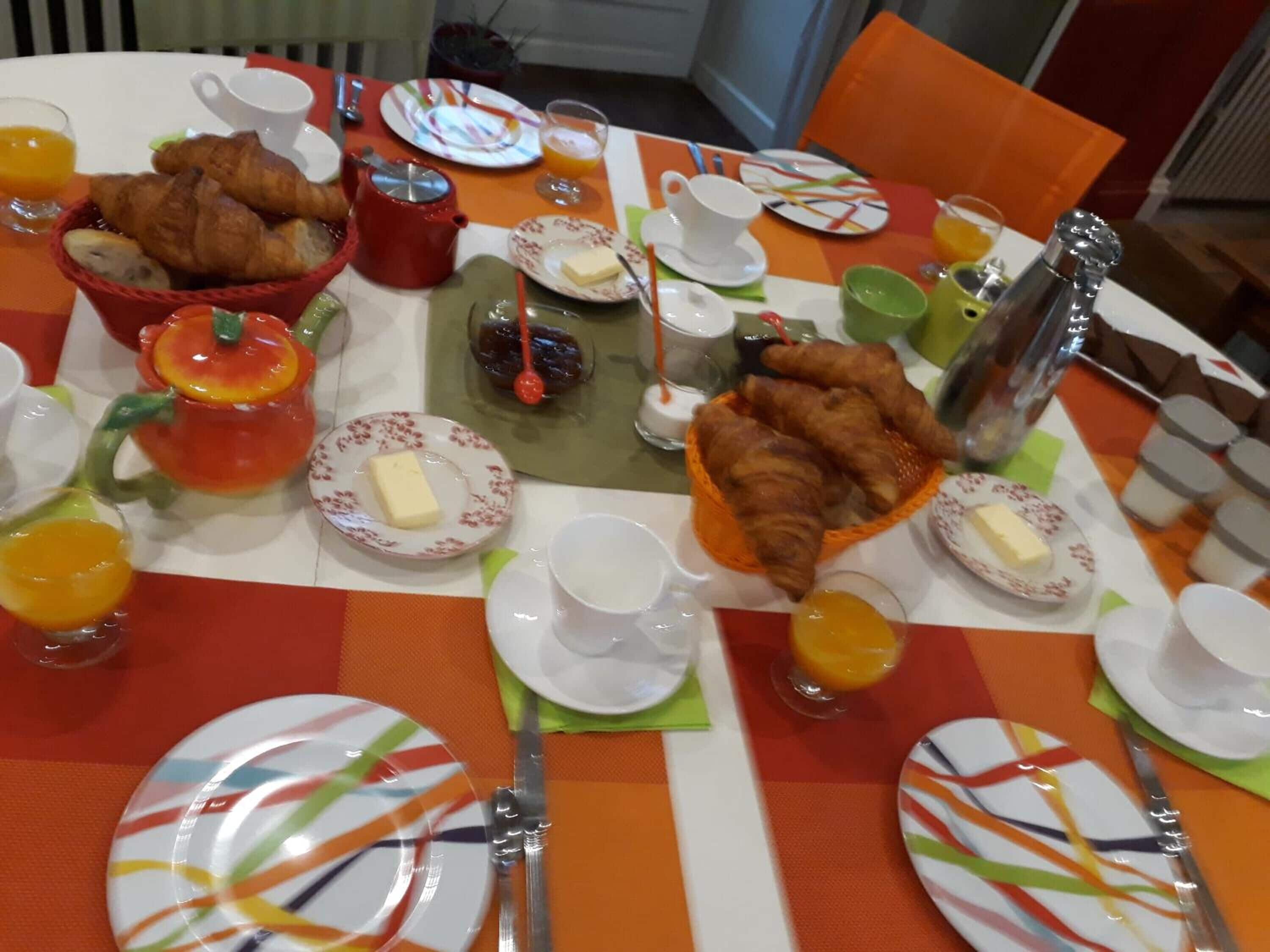 A breakfast setting with croissants, orange juice, and a teapot on a table with colorful plates and napkins.
