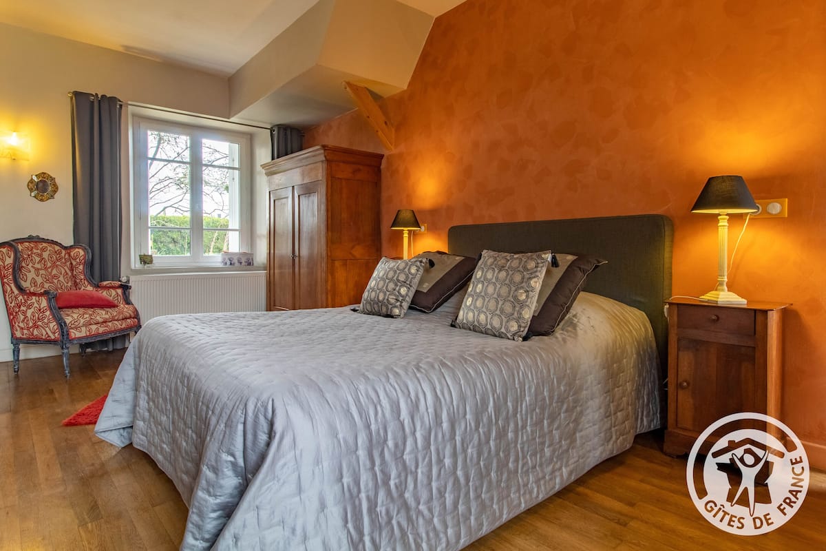 Une chambre à coucher avec un grand lit, deux lampes de chevet, une armoire en bois, un fauteuil rouge et une fenêtre avec des rideaux.