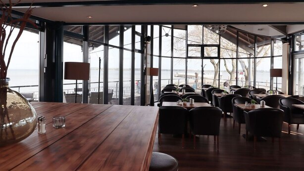 A restaurant with a wooden table and chairs, large windows, and a view of the sea. - Hotel Utkiek (Greifswald)