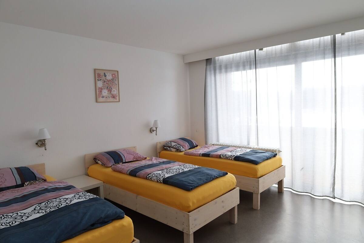 A room with three beds, each with a distinct patterned bedspread, two wall-mounted lamps, and a large window with sheer curtains.
