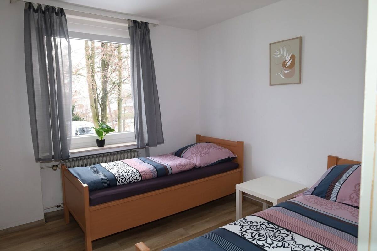 A single bed with a patterned bedspread, a bedside table, a window with curtains, and a radiator.