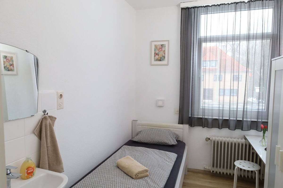 A small, single-bed room with a window, a radiator, a sink, and a mirror.