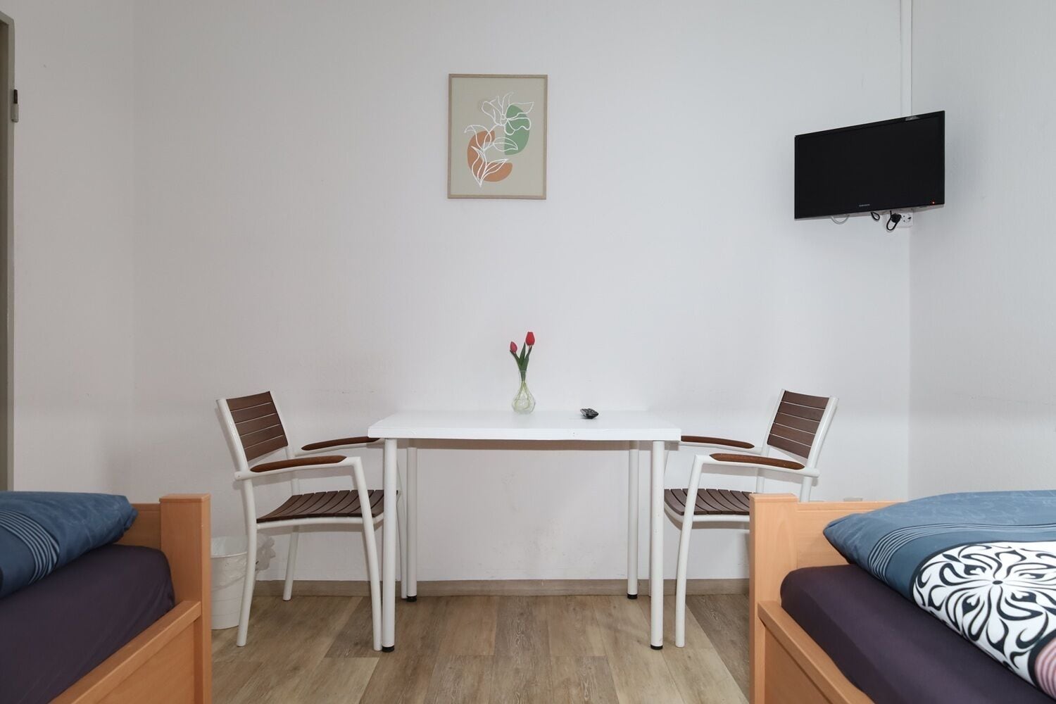 A room with a white table, two chairs, a vase with flowers, a flat-screen TV, and two beds with patterned pillows.