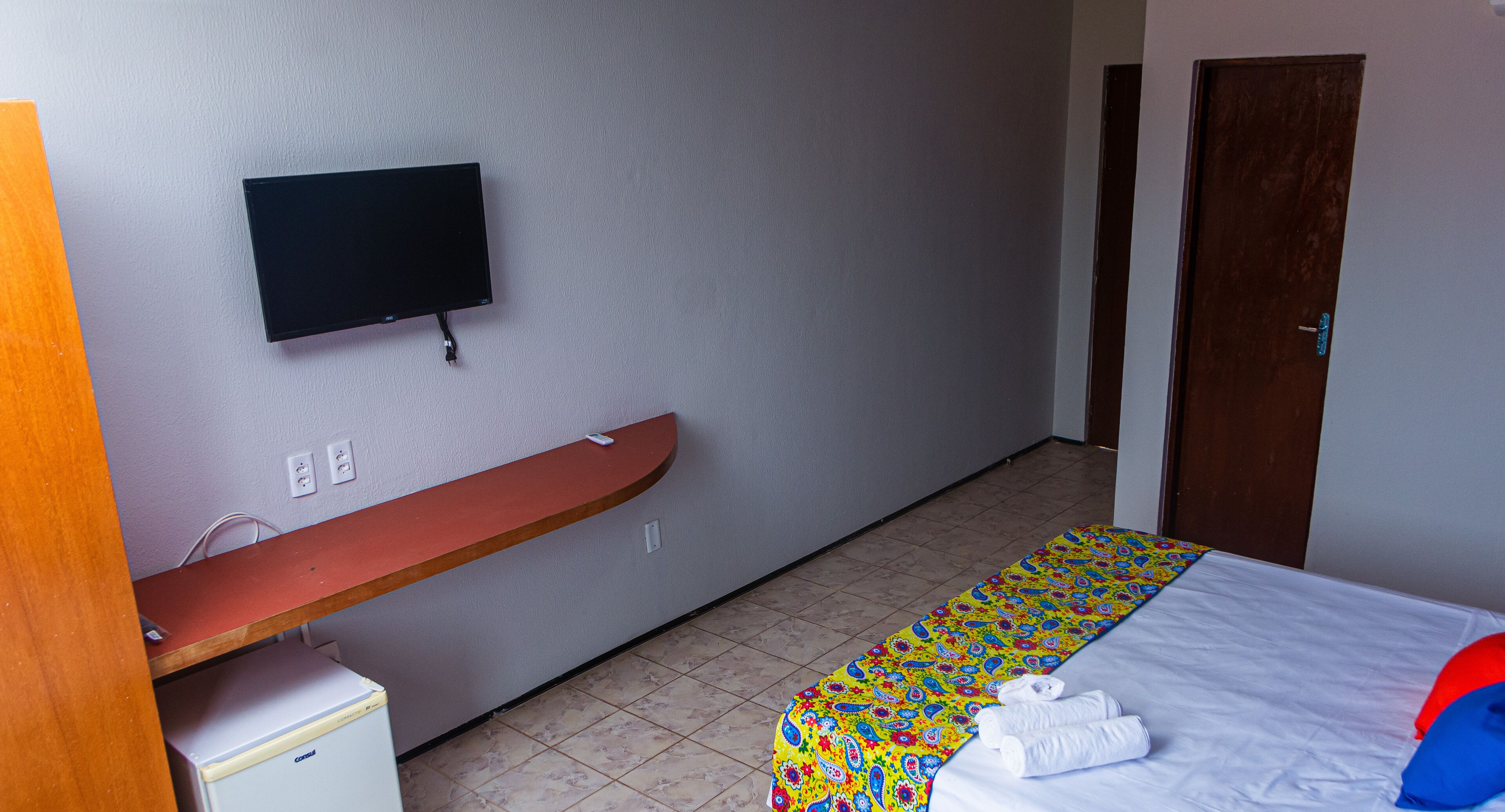 A hotel room with a bed, a TV mounted on the wall, a wooden bench, a small refrigerator, and a door.