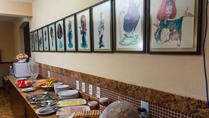 A long buffet table with various food items and a wall of framed caricature artwork. - Kalamari Beach Hotel (Aquiraz)