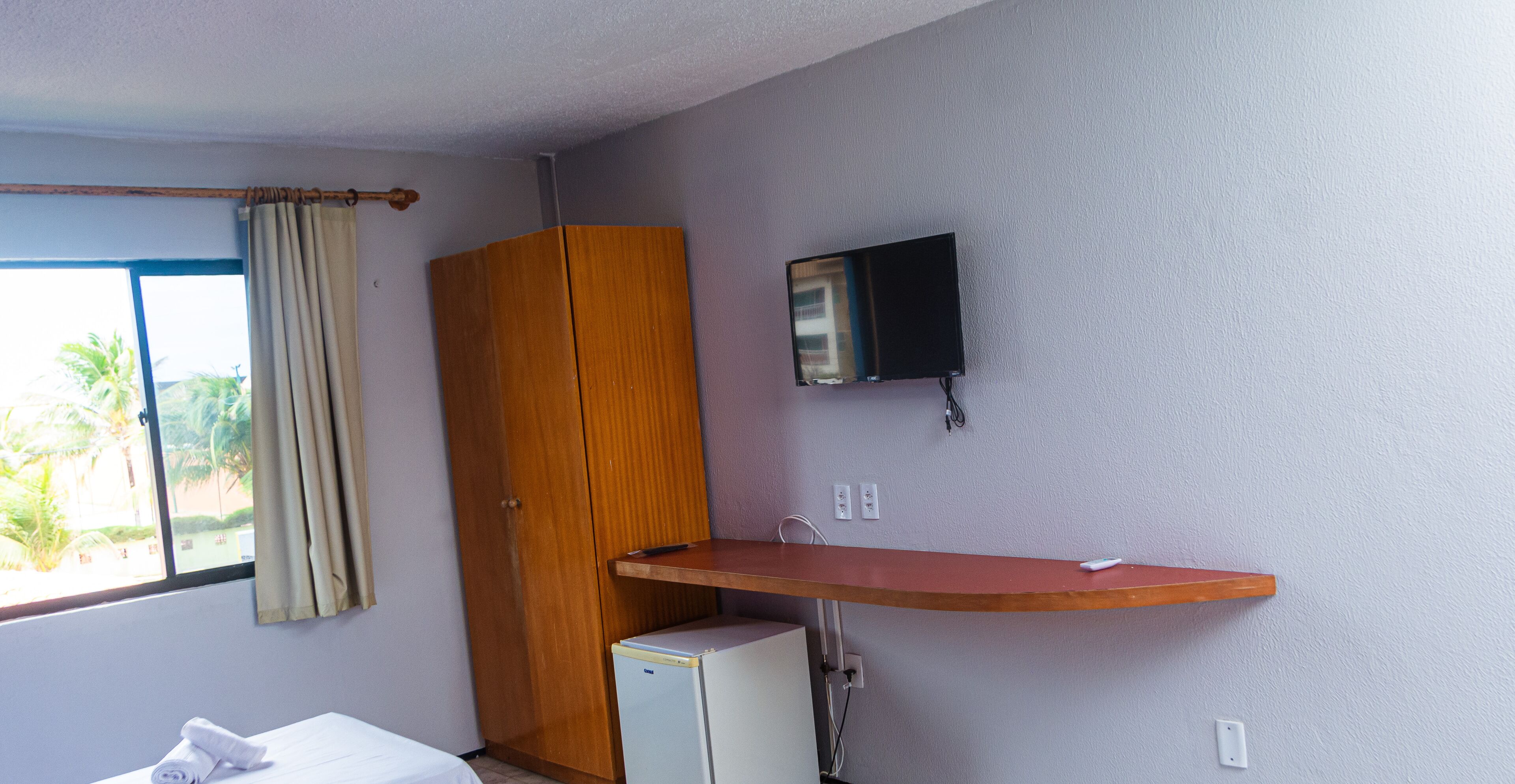 Ein Zimmer mit einem Bett, einem kleinen Kühlschrank, einem Holzschrank, einem an der Wand montierten Fernseher und einem fenster mit Vorhängen.