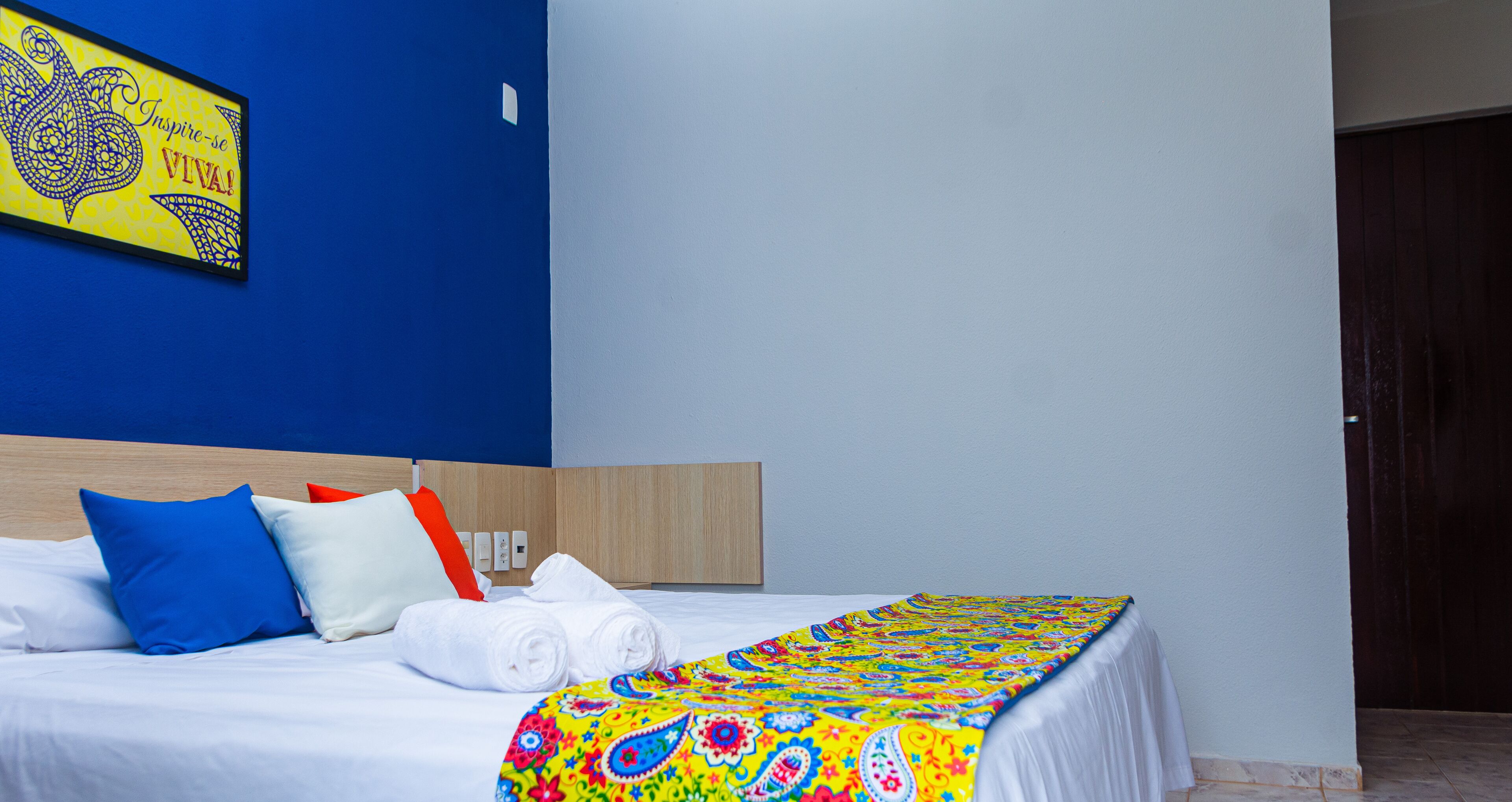 Habitación de hotel con una cama, almohadas, una colcha colorida, una pared azul y un cabecero de madera.