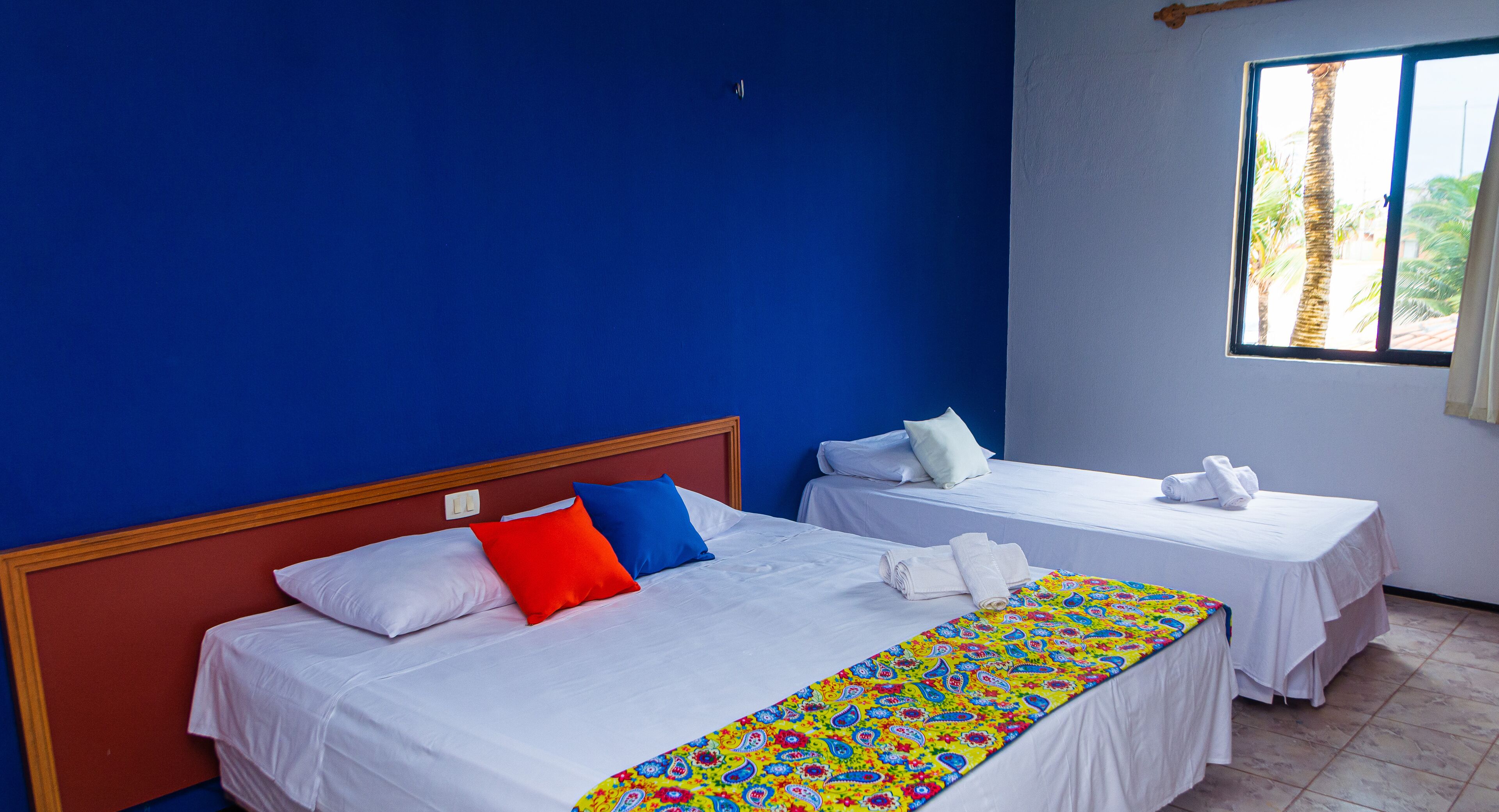 Una habitació d'hotel amb dos llits, un llençol de llit colorit i una finestra que mostra palmeres.