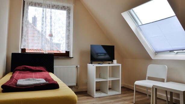A bedroom with a bed, a TV on a stand, a chair, and a window with a view of buildings. - Pension Highway Bünde (Buende)