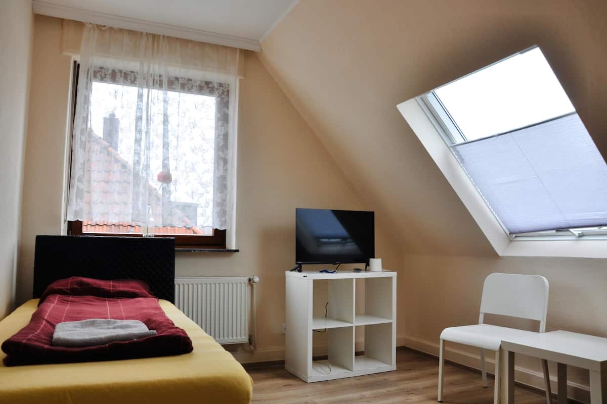 Ein Schlafzimmer mit einem Bett, einem Fernseher auf einem Schrank, einem Sessel und einem Fenster mit Blick auf Gebäude.