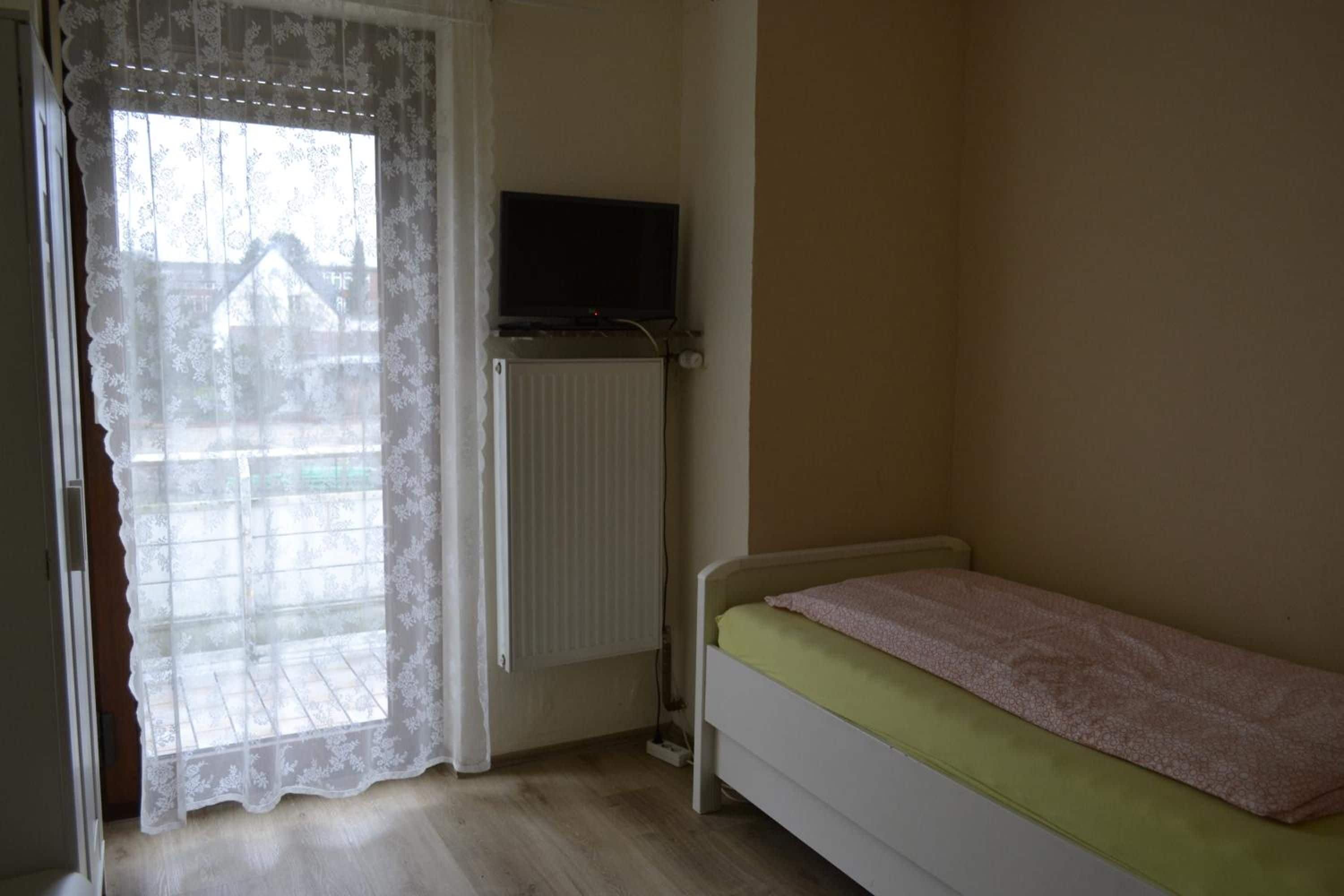 A bedroom with a single bed, a radiator, a television on a stand, and a window with a lace curtain.