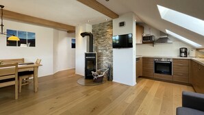 A modern kitchen with a stone wall, wooden beams, a built-in stove, and a dining area with a table and chairs. - Biohof-Rechenmacher (Uffing am Steffelsee)