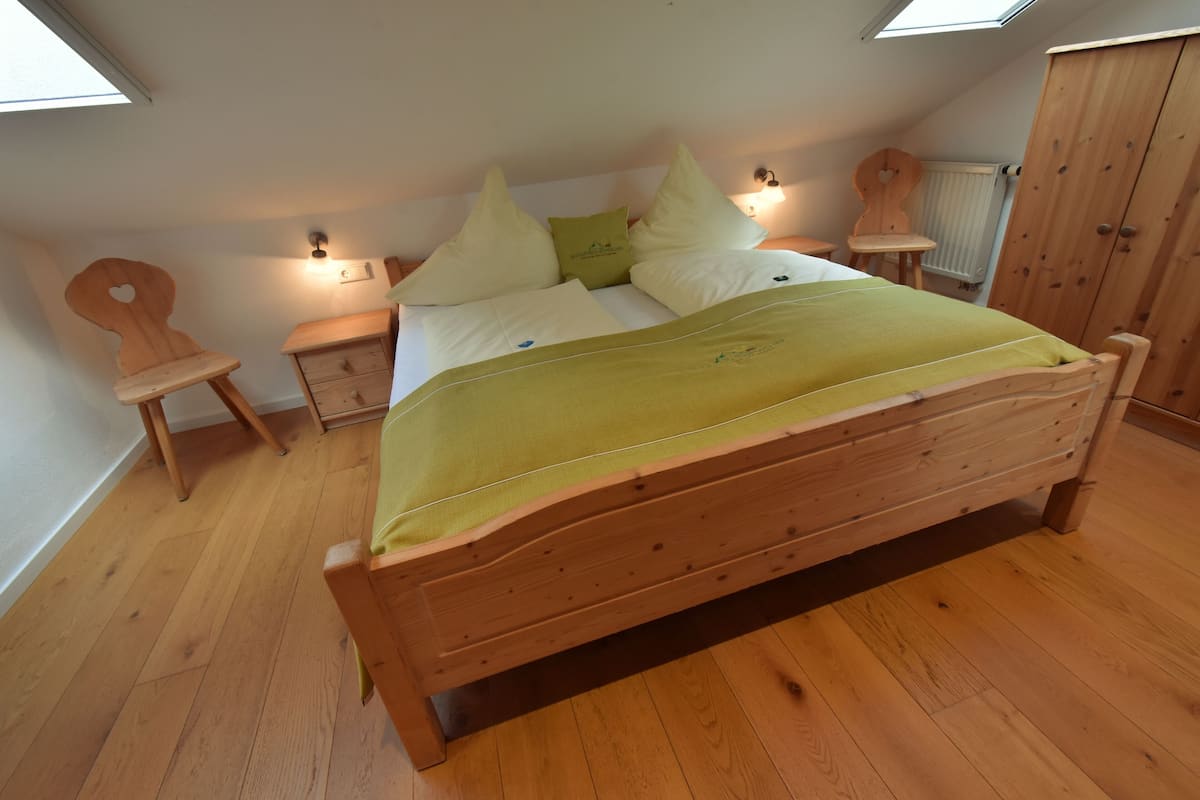 A wooden bed in a room with a skylight, a wooden chair, a small bedside table, and a wardrobe.
