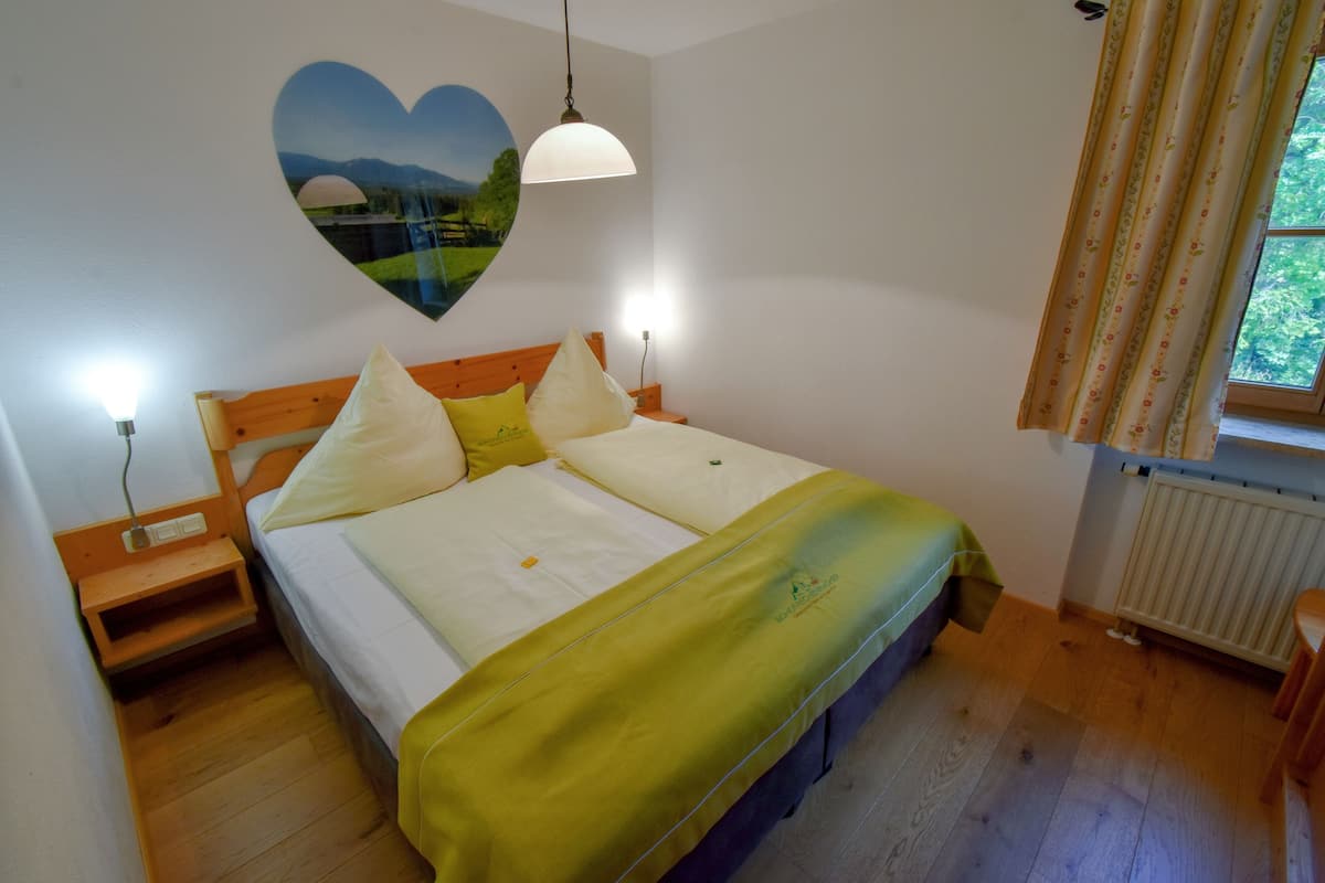 A bedroom with a bed, bedside tables, a nightstand, a wall-mounted heart-shaped decoration, a window with curtains, and a radiator.