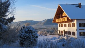 A white building with a sloped roof and wooden balcony, surrounded by snow-covered trees and a mountainous landscape. - Biohof-Rechenmacher (Uffing am Steffelsee)