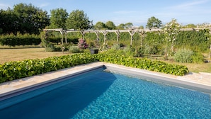 Una piscina azul cristalina con borde de piedra, rodeada de abundante follaje y una pérgola de madera.