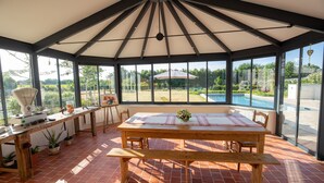 Un porche cubierto con una mesa de madera y bancos, ventanas grandes y vistas a una piscina.