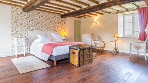 Un dormitorio con techo de madera, papel tapiz floral, una cama con ropa de cama blanca, un baúl y una silla con tapicería floral.