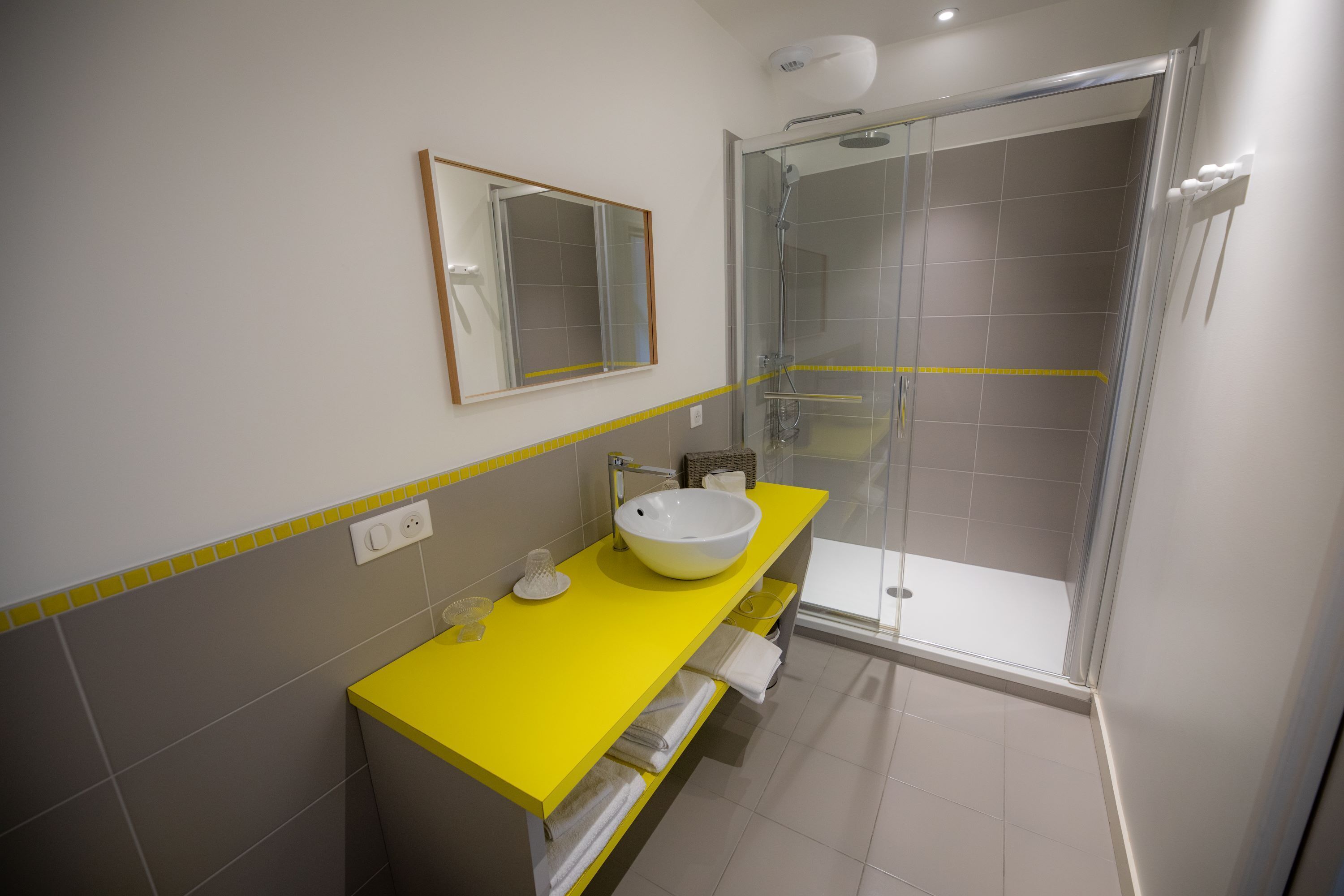 Een moderne badkamer met een geel aanrecht, een witte wastafel en een douche met glazen wanden.