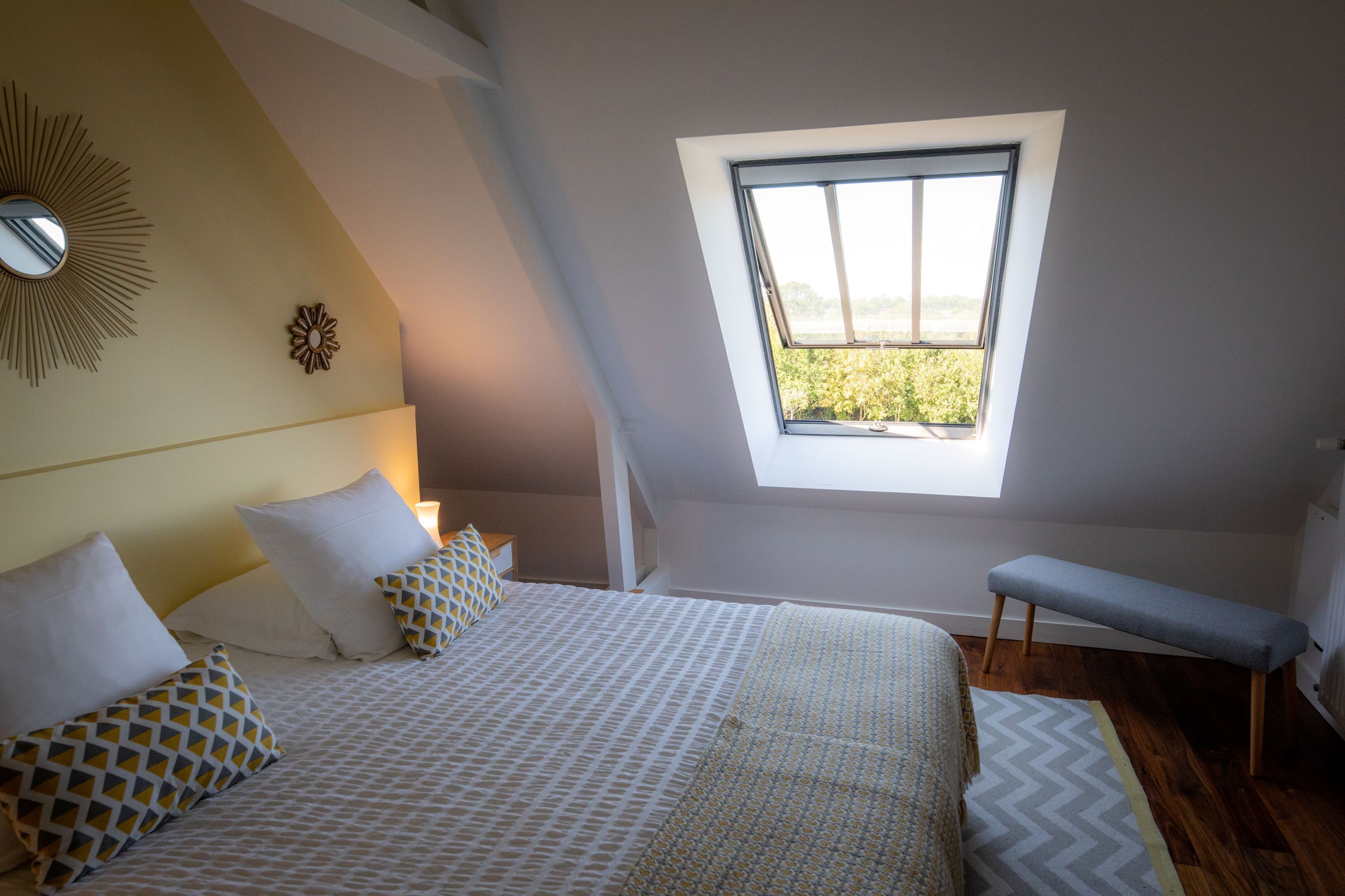 A bedroom with a bed, a bench, a skylight, and a view of greenery outside.