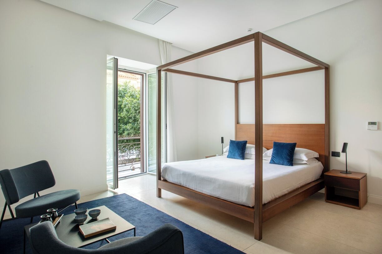A modern bedroom with a four-poster bed, a desk with a chair, and a view of the outdoors through a sliding glass door.