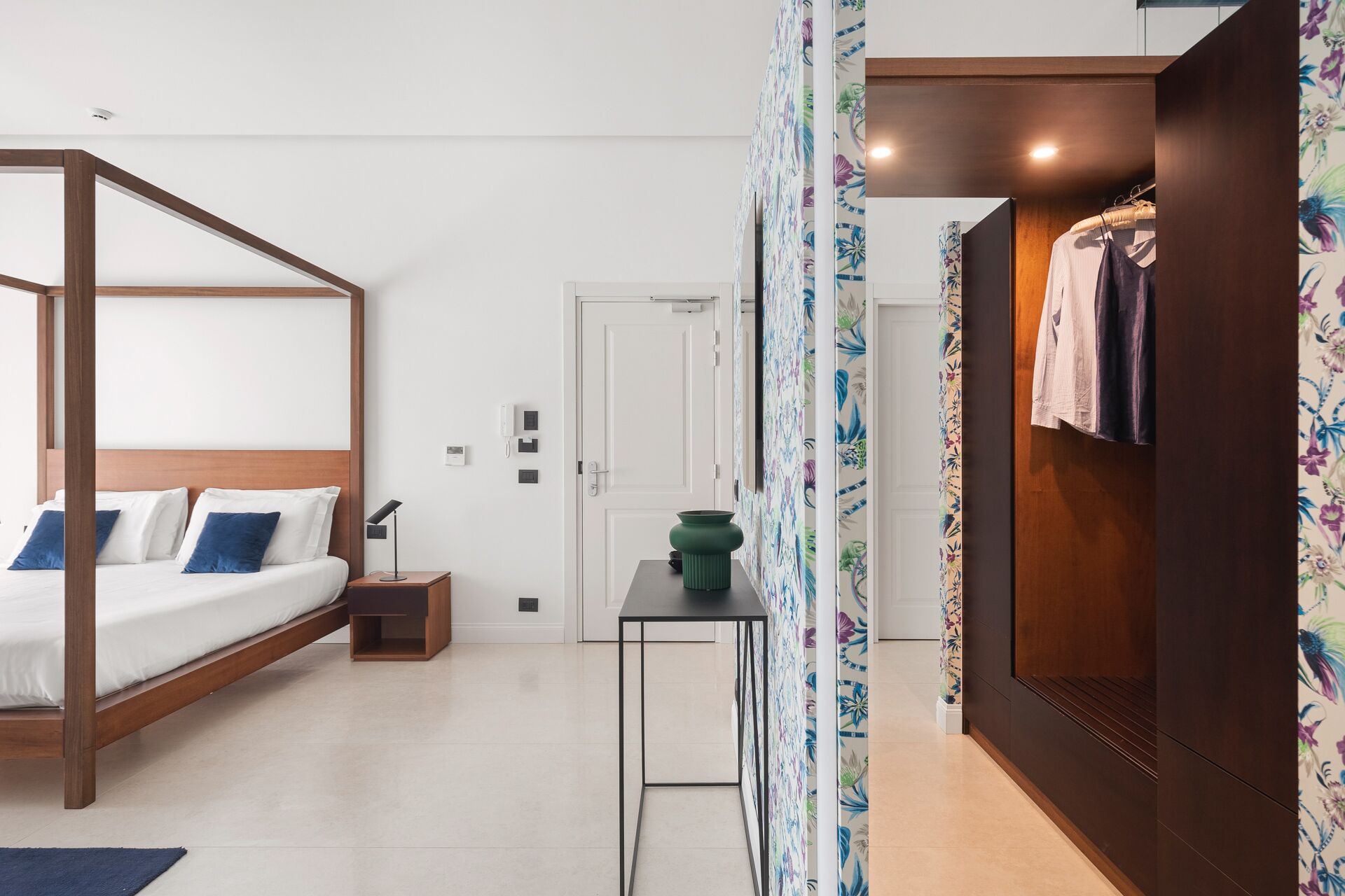 Ein Schlafzimmer mit Himmelbett, Nachttisch mit Lampe, kleiner Tisch mit Vase und Kleiderschrank mit Kleidung.