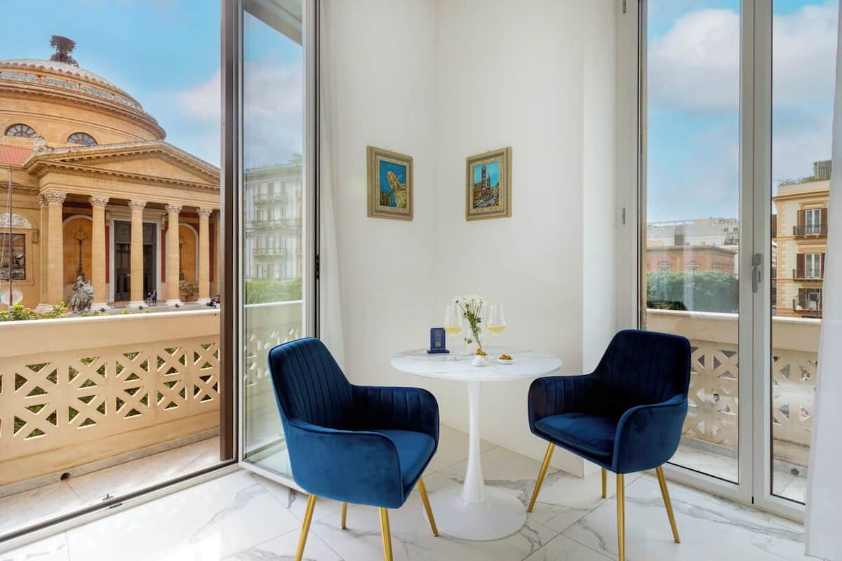 A balcony with two blue chairs, a small table with a vase and glasses, and two framed pictures on the wall.