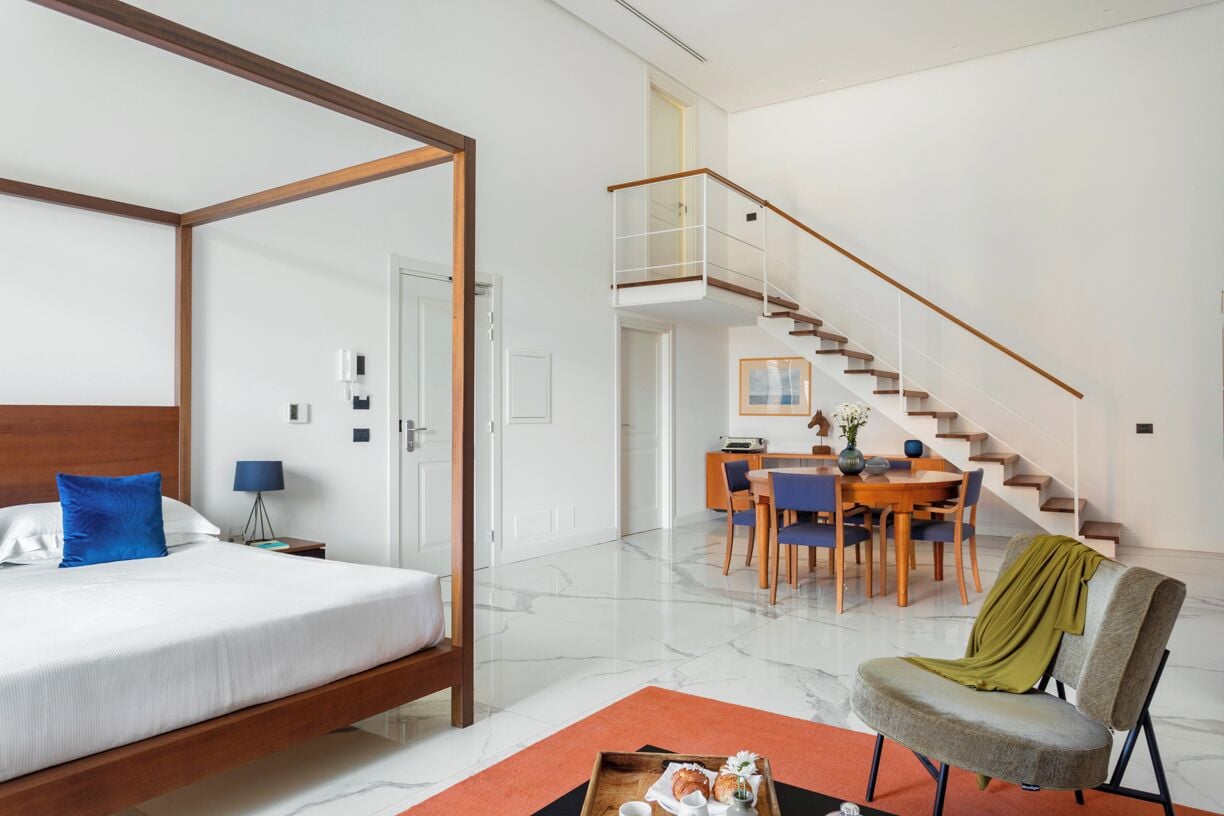 Una habitación de hotel moderna con una cama con dosel, una zona de comedor con una mesa y sillas de madera, y una escalera que conduce a un nivel superior.