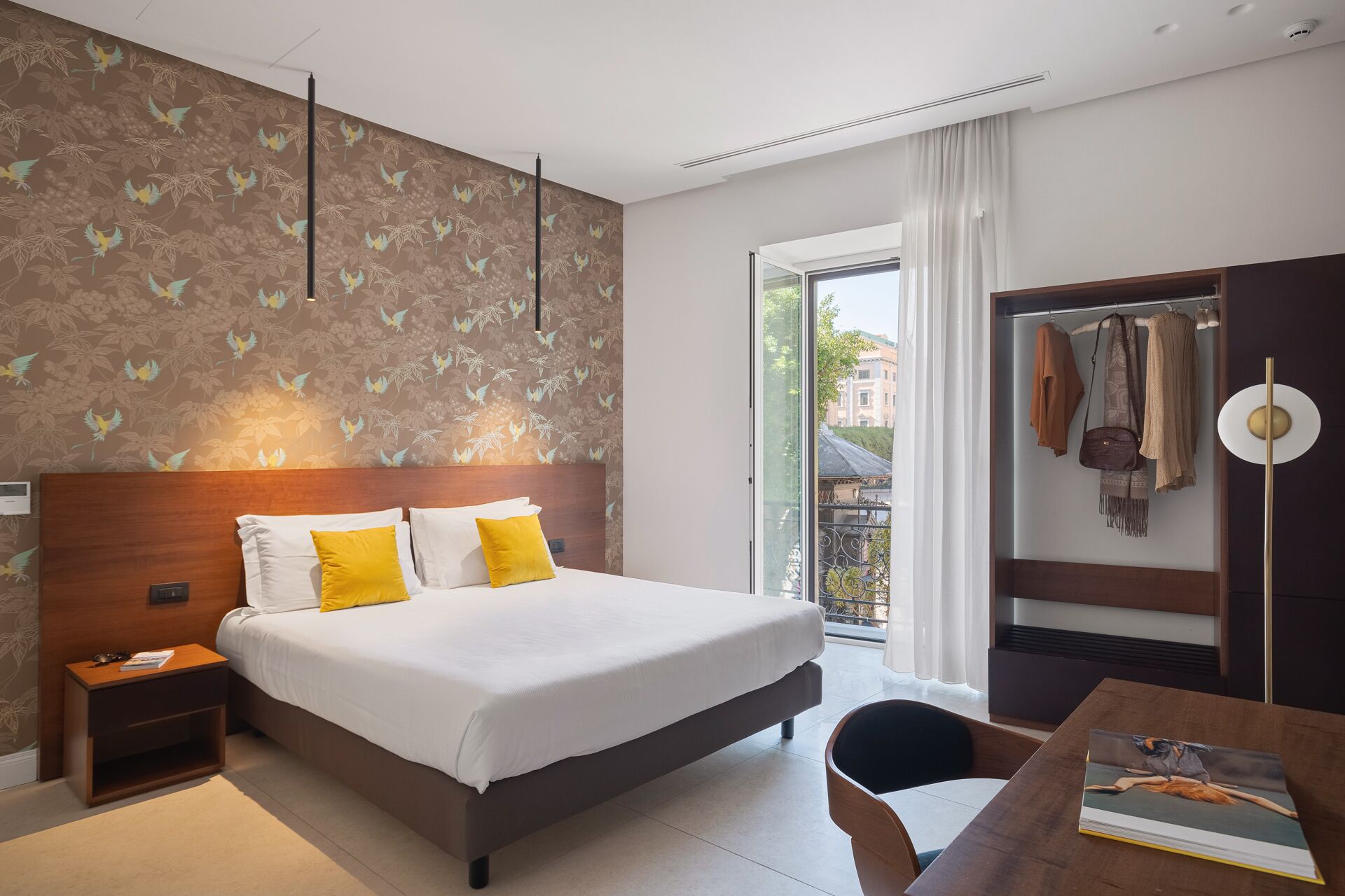 Ein Hotelzimmer mit einem großen Bett, einem Nachttisch, einem Schreibtisch mit Stuhl, einem Kleiderschrank und einem Fenster mit Blick auf Gebäude.