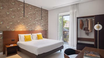 Ein Hotelzimmer mit einem großen Bett, einem Nachttisch, einem Schreibtisch mit Stuhl, einem Kleiderschrank und einem Fenster mit Blick auf Gebäude.
