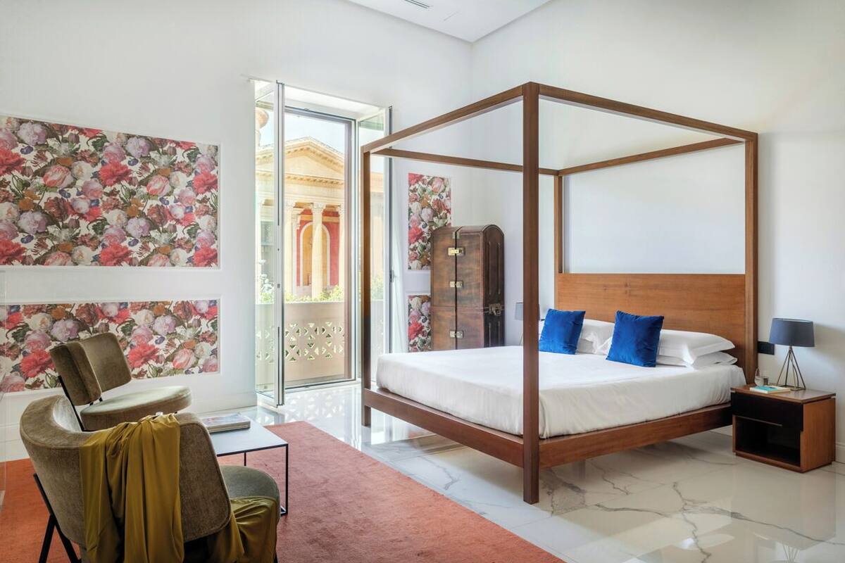 A modern bedroom with a four-poster bed, a chair, a nightstand, and a large window with a view of a building.