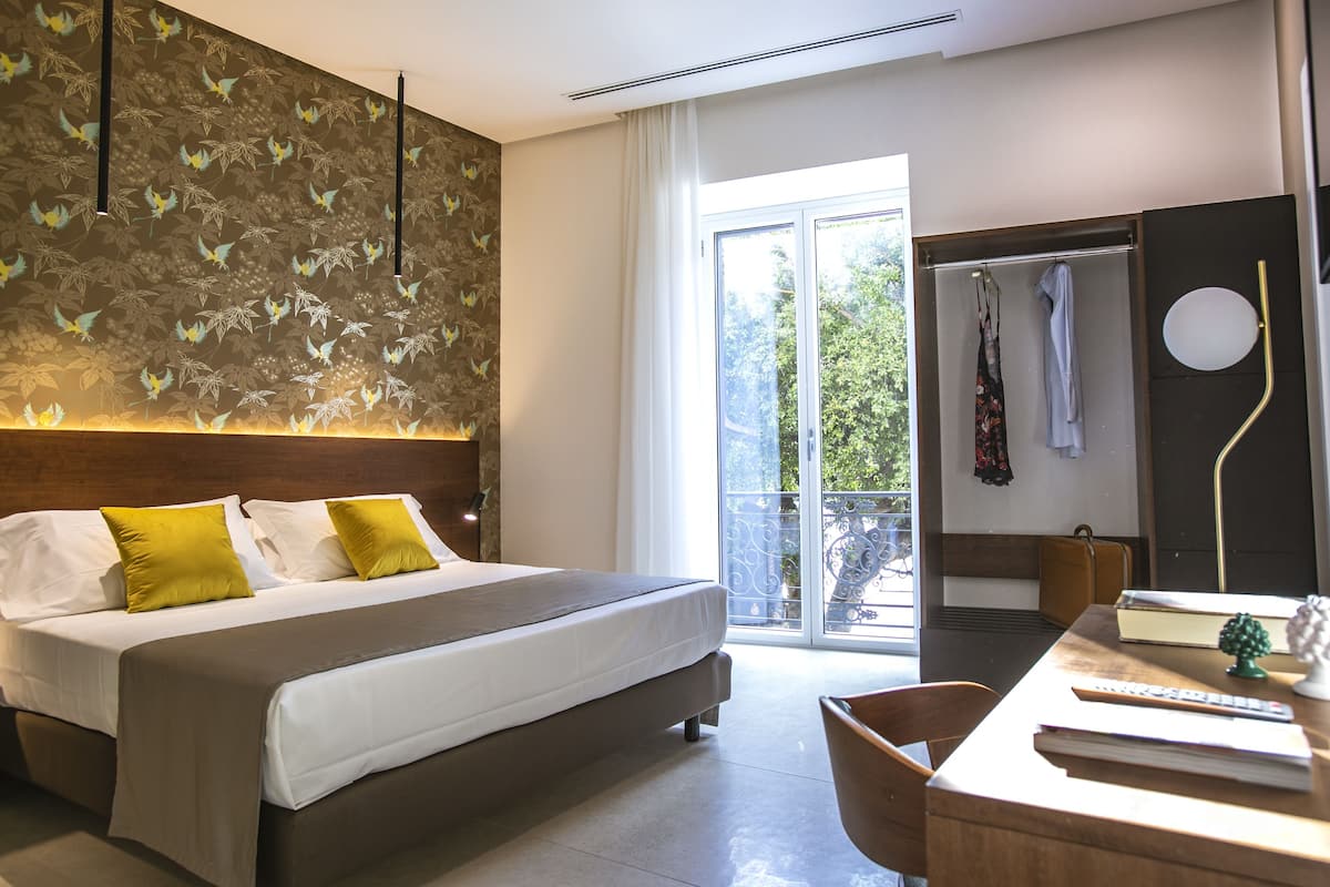 A modern hotel room with a large bed, a desk with a chair, a balcony with a view, and a shower area.