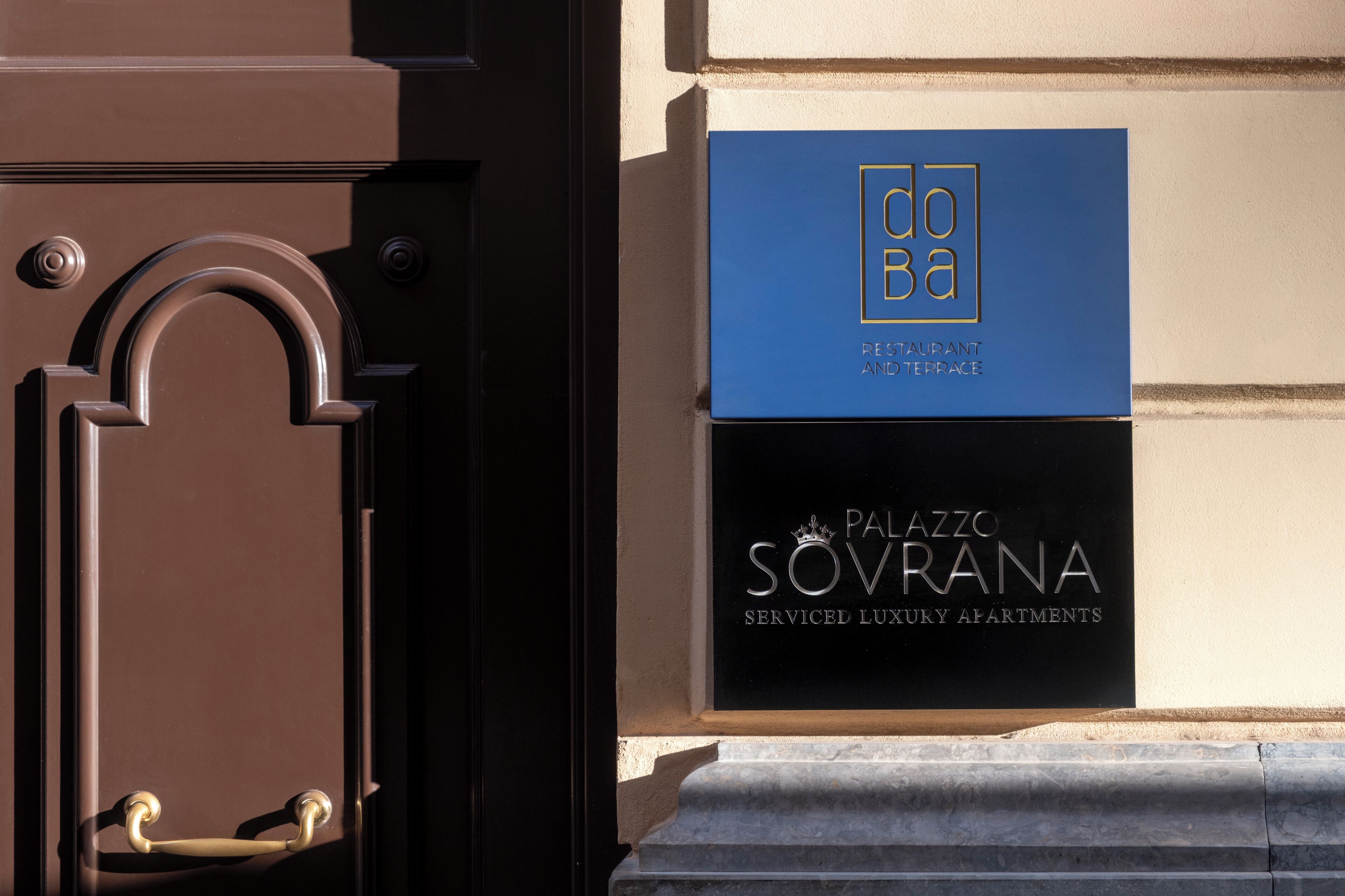 Una puerta con manija de latón y un buzón con dos letreros: "DOBA" y "PALAZZO SOVRANA SERVICED LUXURY APARTMENTS".