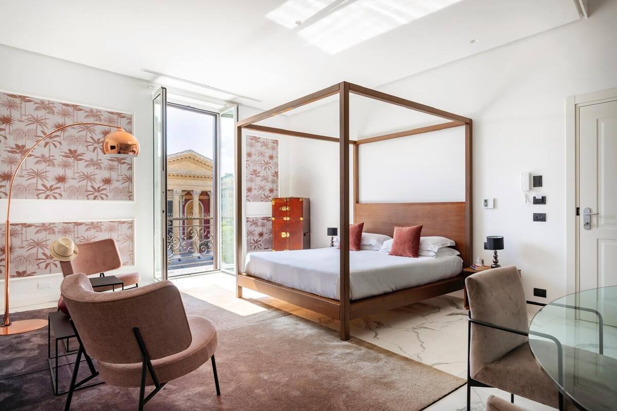 A modern bedroom with a four-poster bed, a glass table, and a view of a classical building through a glass door.