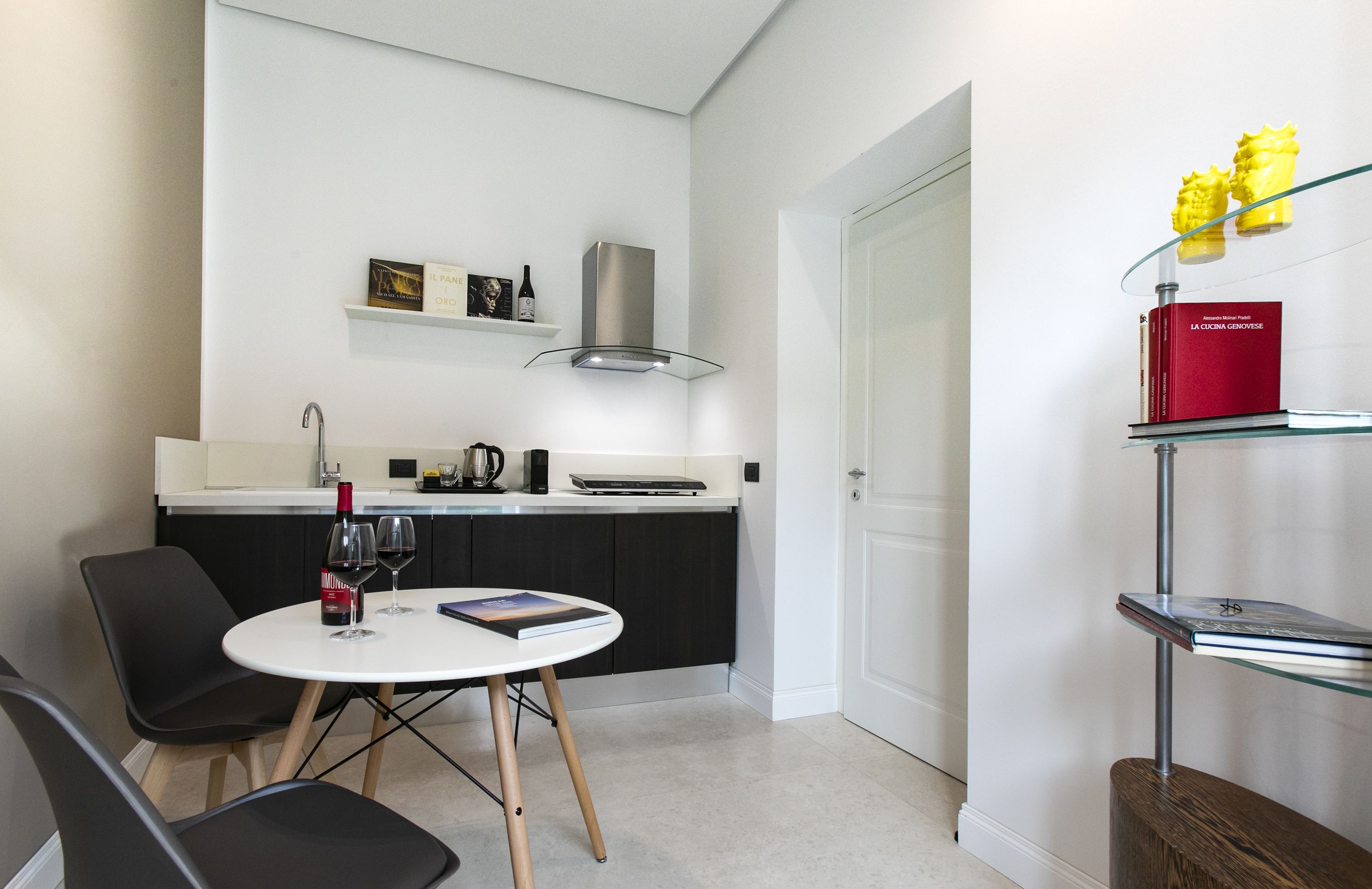 A modern kitchen with a white countertop, black cabinets, and a stainless steel range hood. There is a dining area with a round table and chairs, and a bookshelf with books and decorative items.
