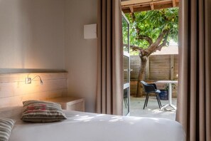A bedroom with a bed, a bedside table, a lamp, and a view of an outdoor area with a table and chairs. - Hôtel de L'Océan (Moliets-et-Maa)